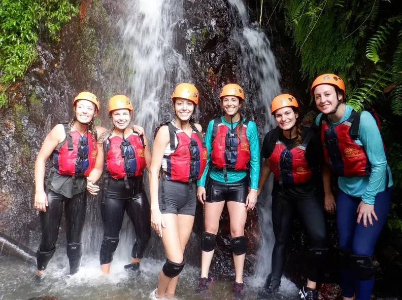 Guided group of six guests in helmets and life jackets standing under a rainforest waterfall